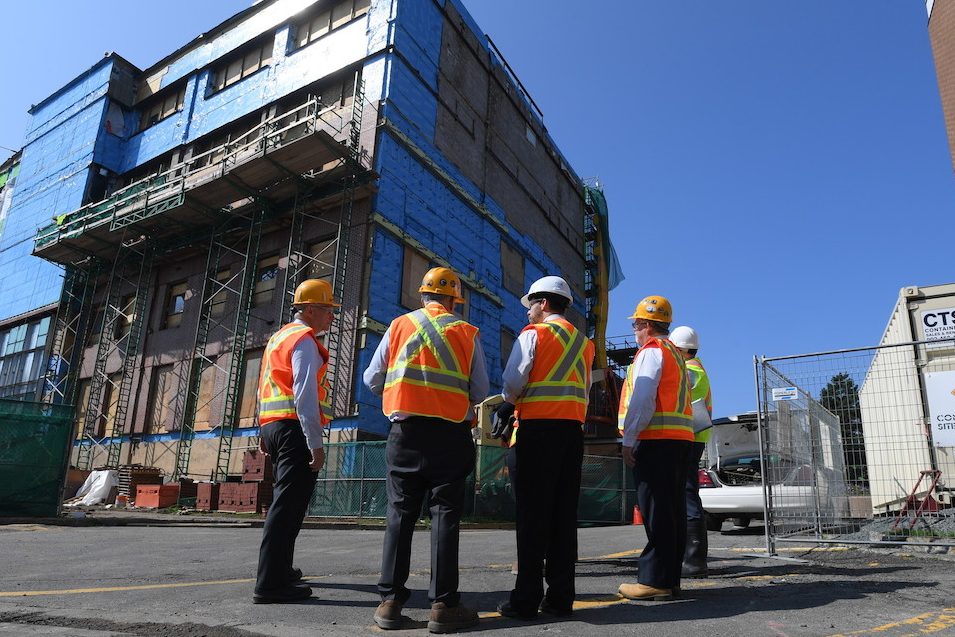 Four workers wearing hard hats and safety vests viewed from behind in front of the IDEA Building while it was under construction.