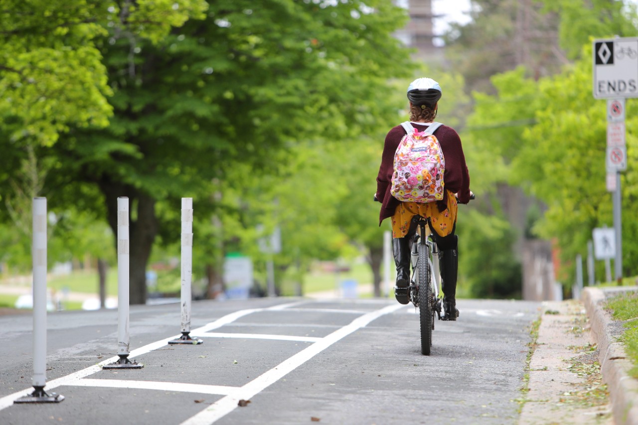 A woman wearing a helmet and colourful backpack cycles in a protected bike lane on a tree-lined street.