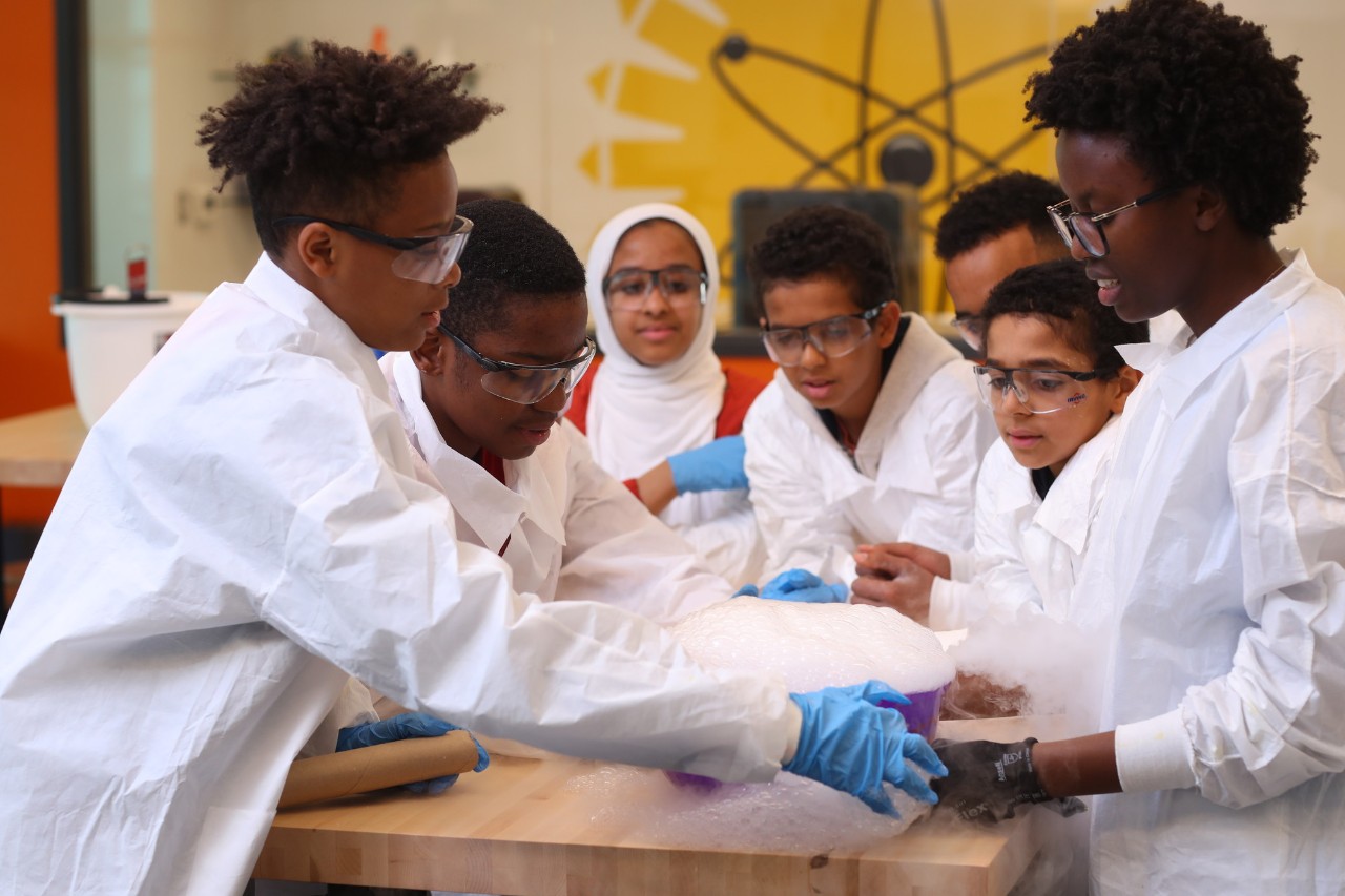 A group of students from diverse backgrounds wear white lab coats while they conduct a science experiment at Dalhousie's Imhotep Legacy Academy. 