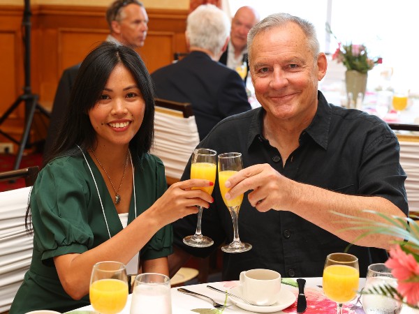 A man and a woman sit at a table, smiling and toasting with glasses of orange juice.