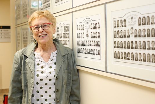 Glenda Butt stands in front of a wall of class photos of dental hygiene alumni. 