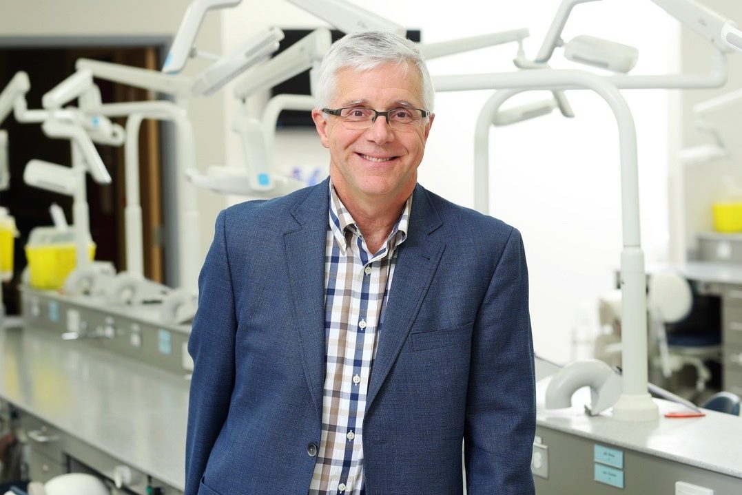 Dr. Lee Erickson stands smiling in a dental lab dressed in business casual attire.