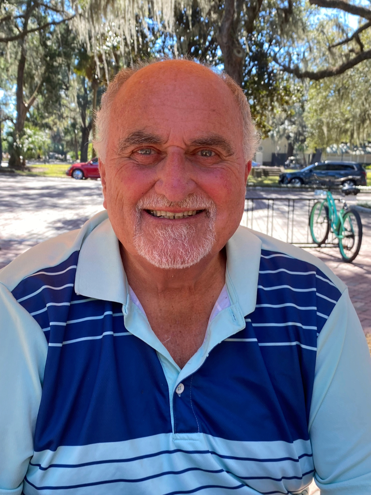A man dressed casually smiles outdoors in fine weather with a tree-lined street and bicycle rack in the background.