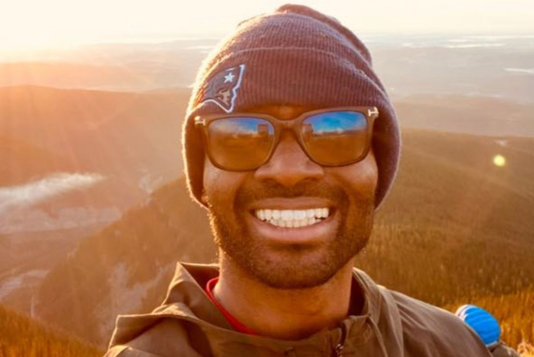 A male-presenting person wearing a toque and sunglasses smiles outdoors with a mountain vista in the background. 