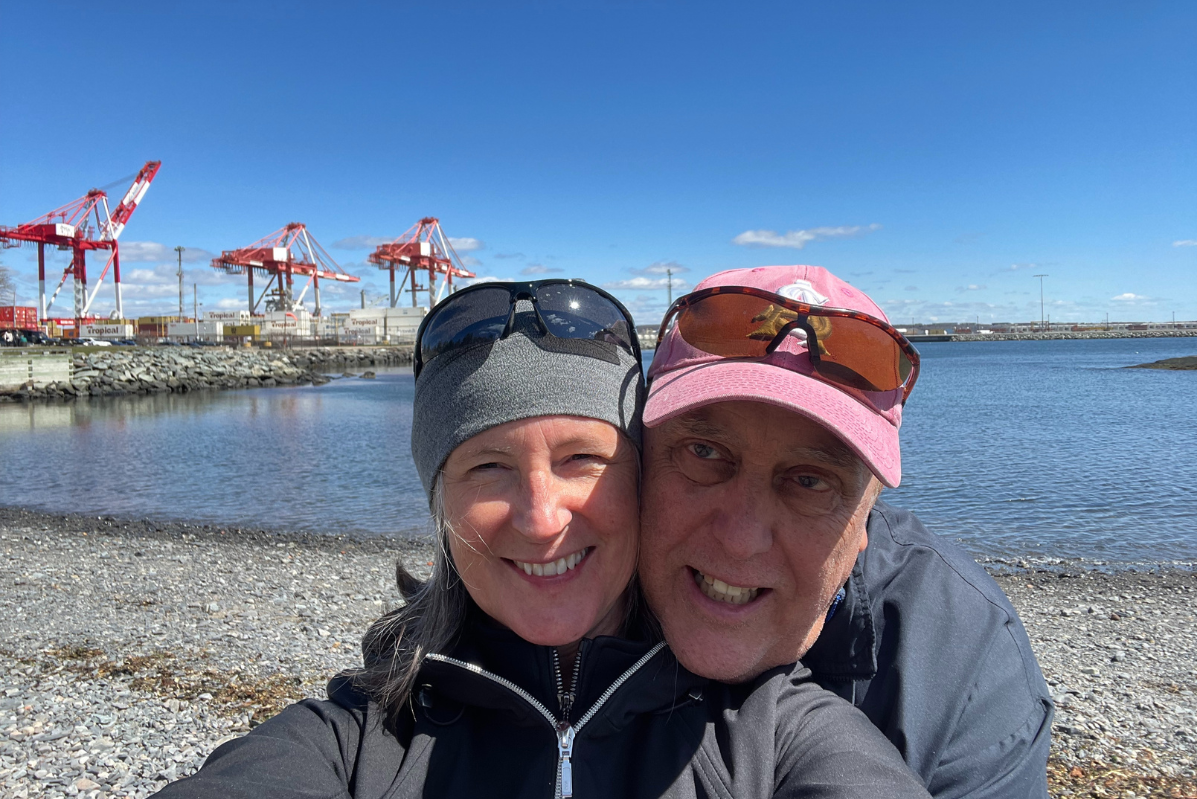 A couple wears sunglasses on their heads and hats. They smile and snuggle for a selfie on the Halifax waterfront. 