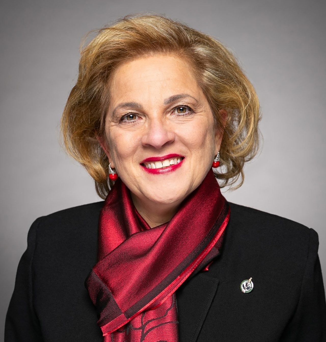 A portrait of a light-skinned woman with short sandy blond hair; she smiles at the camera. She is wearing a red scarf tied at the neck, and matching red lipstick and earrings. Her blazer is black with a silver lapel pin.