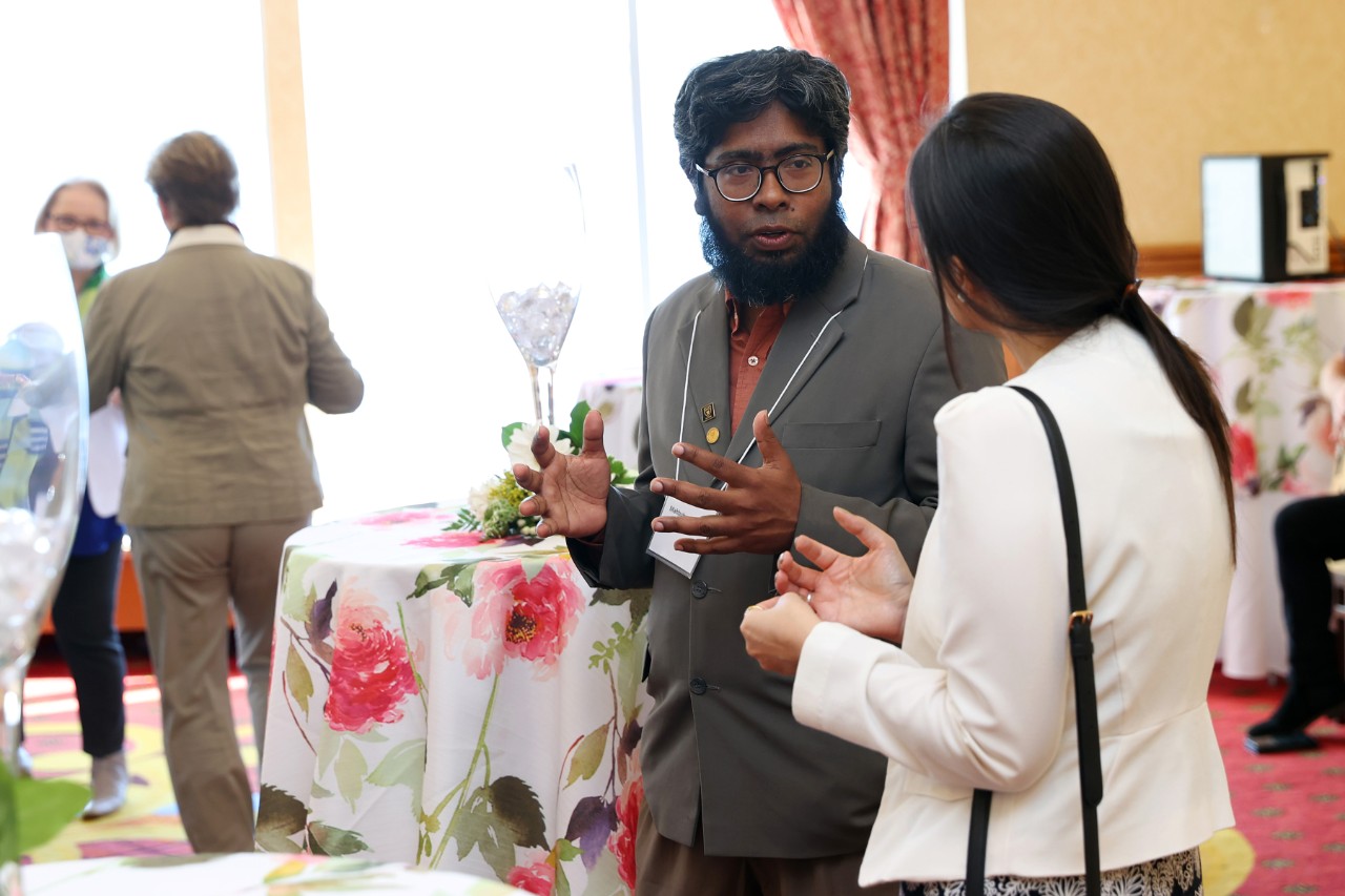 Rahman is standing and in conversation with a woman at the Champagne Social event.