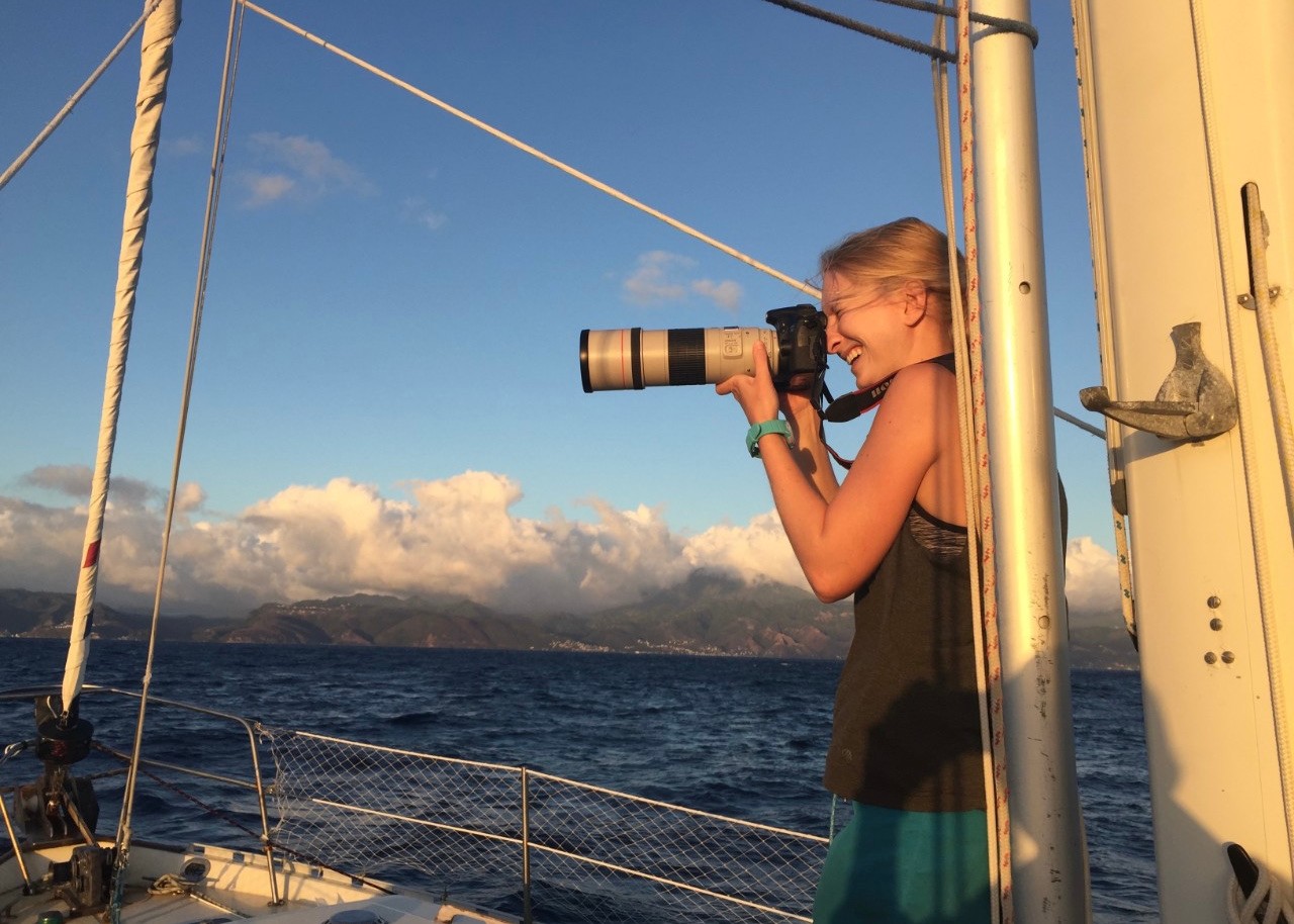 A person with long blonde hair stands on a ship in the ocean with clouds at the horizon taking a photo with a telephoto lens on a camera.