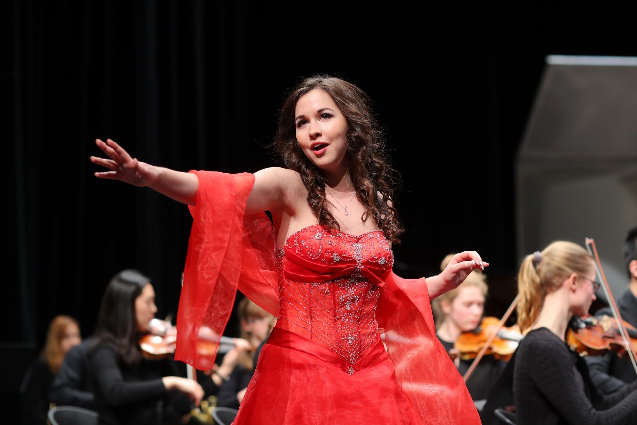 A soprano wears a red ballgown and sings animatedly in front of an orchestra. 