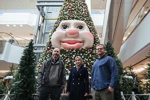 Three people are standing in front of Woody the the Talking Christmas Tree in the atrium of a three-story mall.
