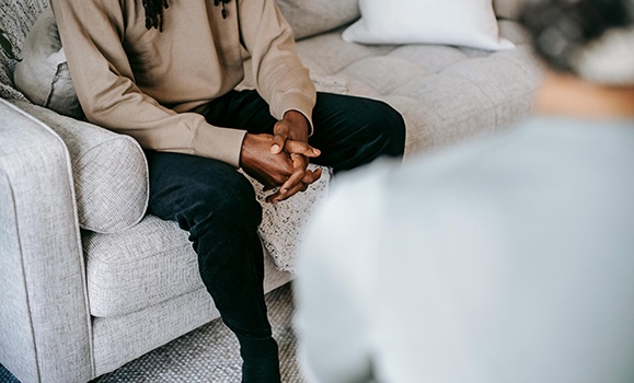 Over a blurry shoulder you can see someone with folded hands sitting on a couch.