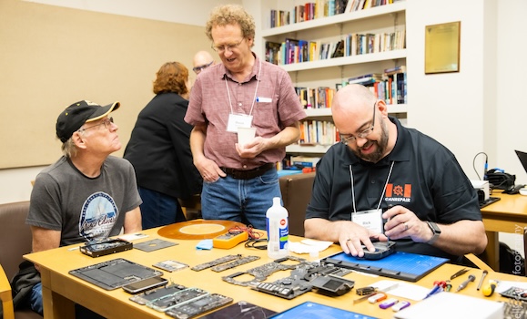 Three men gather around a table. One is fixing a small electronic. One is looking over his shoulder and chatting with the third.