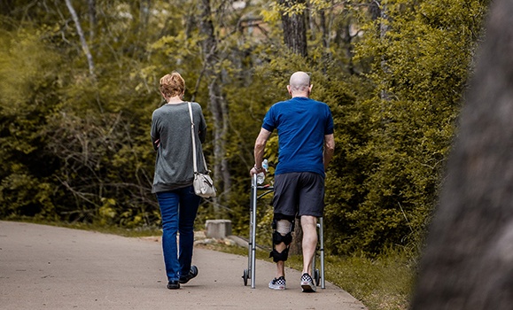 Two people are walking in a park. One is using a walker.