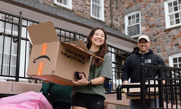 A student is holding a moving box, smiling at the camera. A family member in a baseball cap is holding a box behind them.