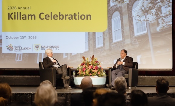 Kim Brooks and Bernie Miller are sitting on on a stage in black chairs with a blue coffee table and flowers between them.