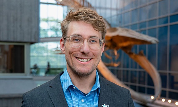 A headshot of Dr. Rob Lennox in the Beaty Centre with the whale skeleton hanging in the background.
