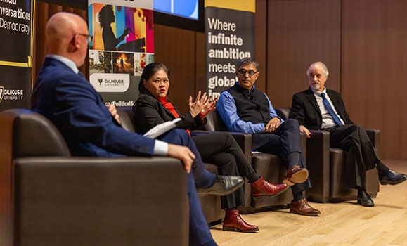 Panelists are sitting on stage in black chairs with pull-up banners behind them.