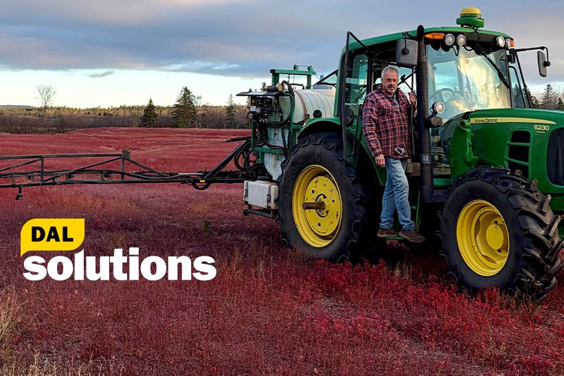 Françoise Baylis is sitting on a beige chair looking at the camera. She is wearing a pink long sleeve top, red and purple scarf, and has black rimmed glasses on.