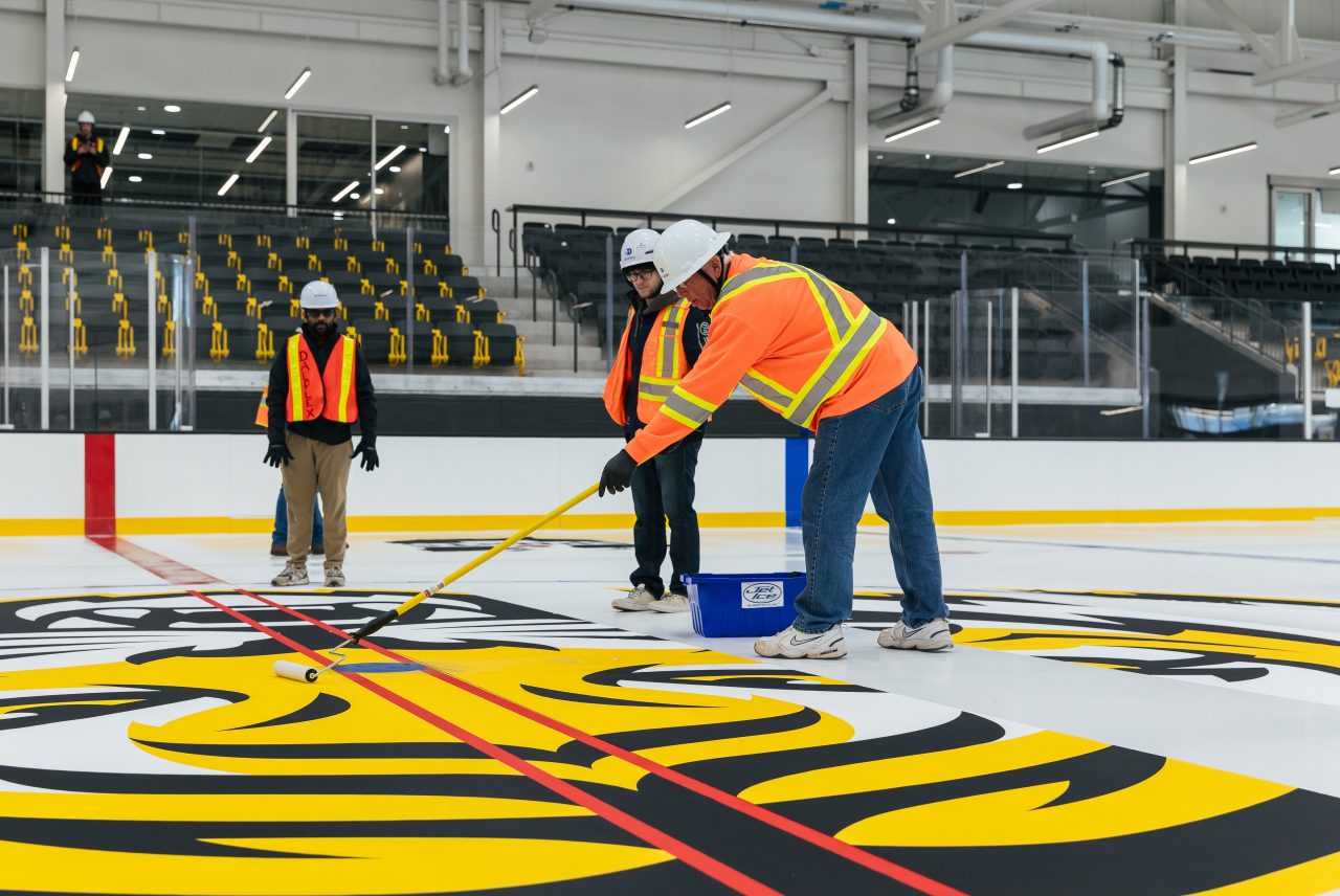 Three staff are wearing orange construction jackets and hardhats are installing a giant Dal Tigers logo at centre ice of a hockey rink.