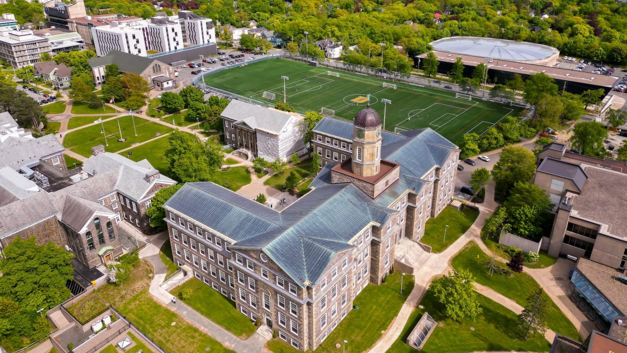 An aerial view of Dalhousie's Studley Campus with an old stone building in the foreground, sports field in the middle, and trees and ocean in the background.