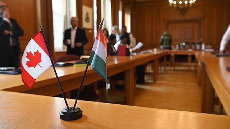 Small, desktop Canadian and Indian flags are displayed on a meeting table while participants network in the background.