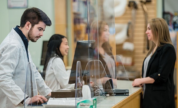 A patient is standing at a pharmacist desk. Two pharmacists are there, both wearing white lab coats.