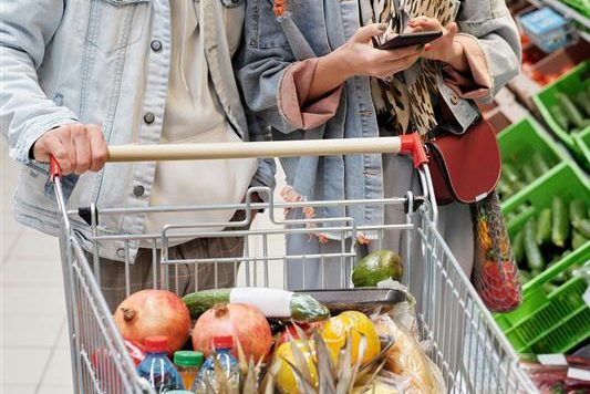 Two people are shopping in a grocery store with a full cart including onions, a cucumber, pineapples, and lemons.