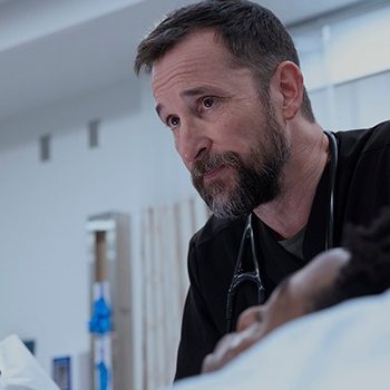 Noah Wyle is wearing black scrubs with a stethoscope looking at a patient off screen.