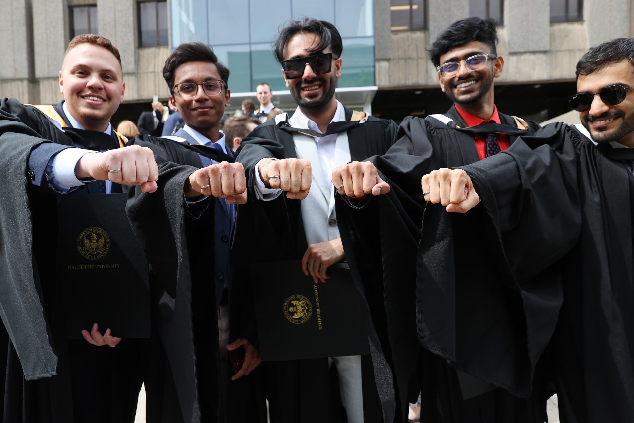 Five graduates are wearing their graduation gowns and holding out their hands in a first to showcase the new iron rings on their pinkeys.
