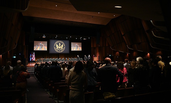 An auditorium is full of families. There are a couple dozen empty chairs on stage. There are three backdrop screens, one with the Dalhousie eagle emblem.