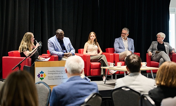 Five panelists are sitting on a stage in red chairs.