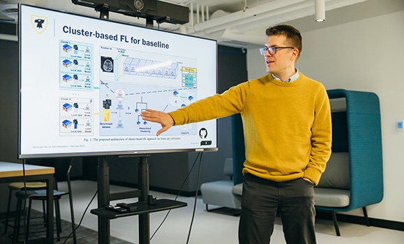 Dr. Frank Rudzicz is wearing a yellow crewneck sweater and standing in front of a screen displaying a Dalhousie logo, graphs, and text.