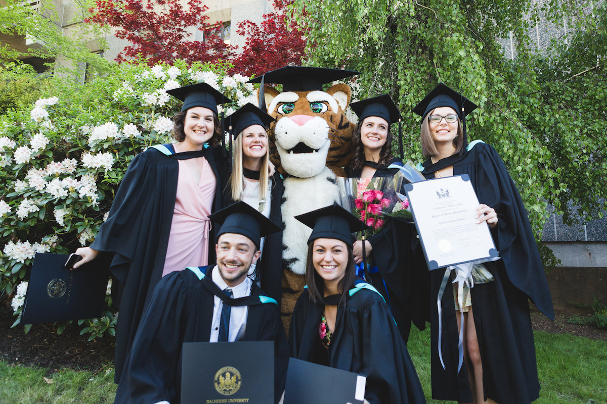 Six students in grad caps and gowns are posting with the Dal Tiger, also in a grad cap, in front of campus foliage. Three students are holding their degrees and one student is holding a bouquet of roses.