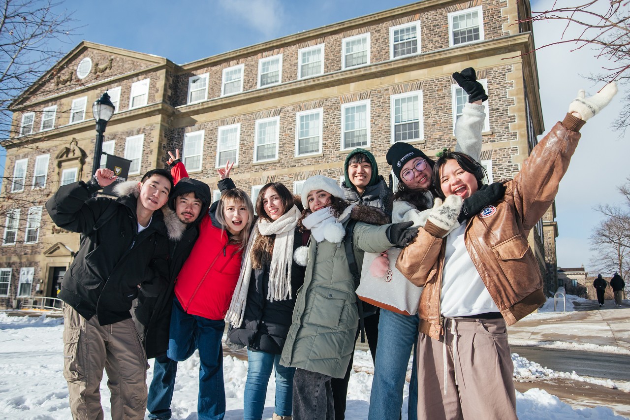 Eight students wearing winter coats, hats, and scarves, smile and wave at the camera on a sunny, snowy day.