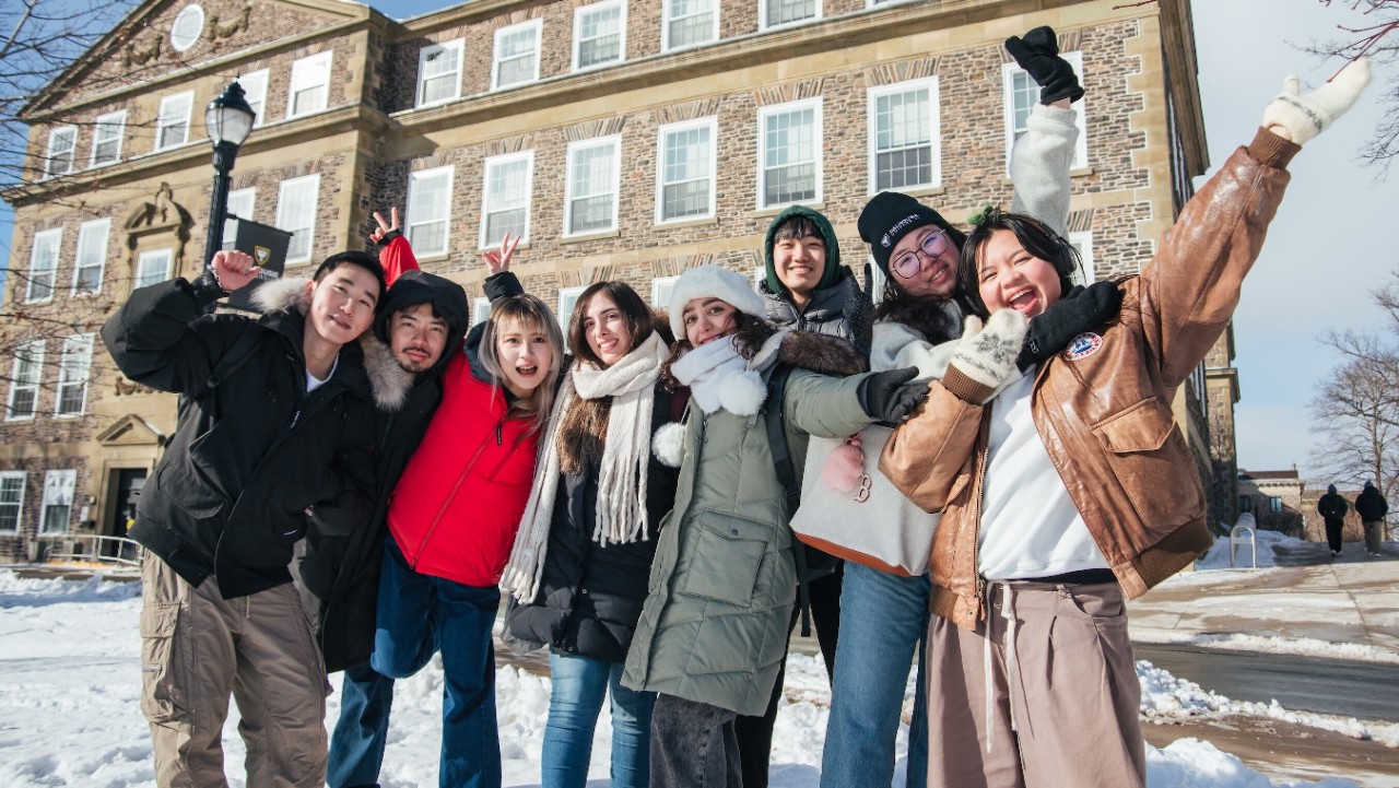 A group of eight students stand on a snow-covered ground and pose for the camera in front of an old stone university building.