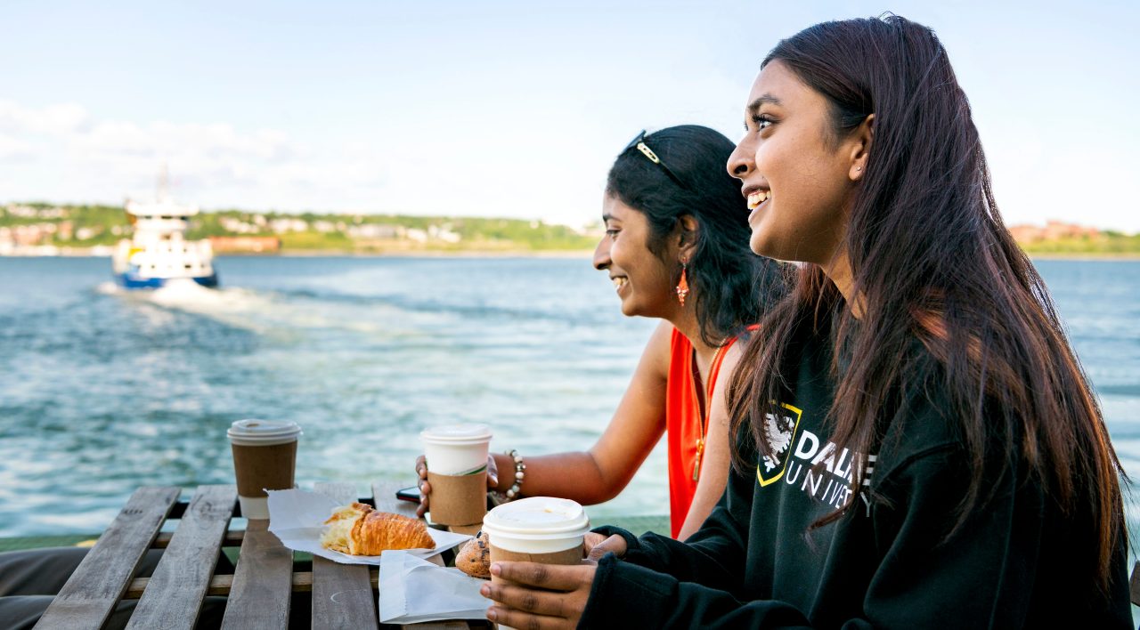 Students sitting at a picnic table with to go coffee cups and croissants. A ferry crosses the harbour in the background.