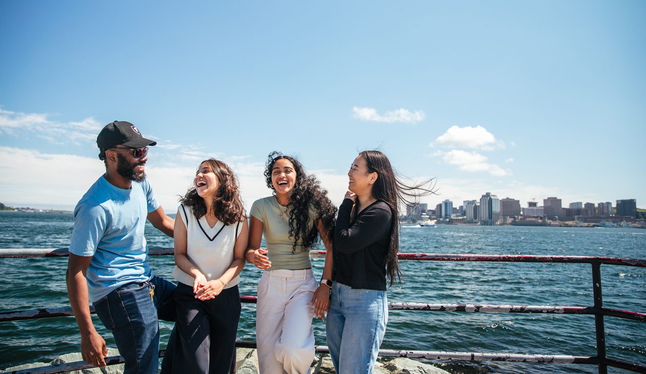 Four students laughing in the sunshine. Behind them is a body of water and a city skyline.