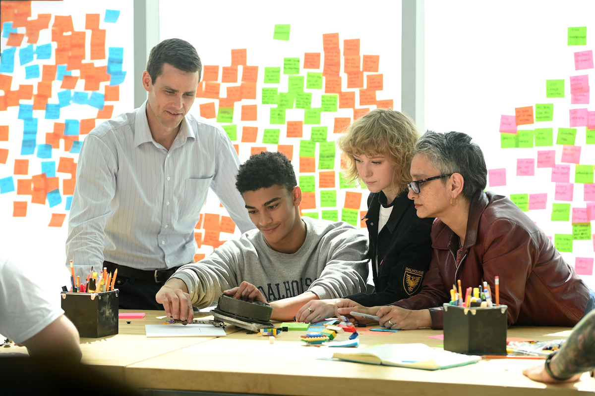 Two students work on an engineering project as their professors look on. Behind them is a wall of colourful post it notes.