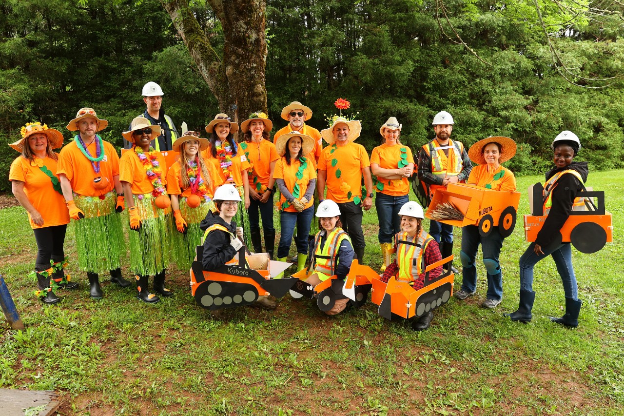 A group of people dressed in orange shirts with fun straw hats and cardboard tractors pose for a photo.