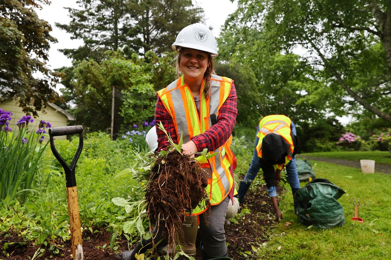 A smiling gardener wearing a hard hat shows off the weeds she has just pulled.