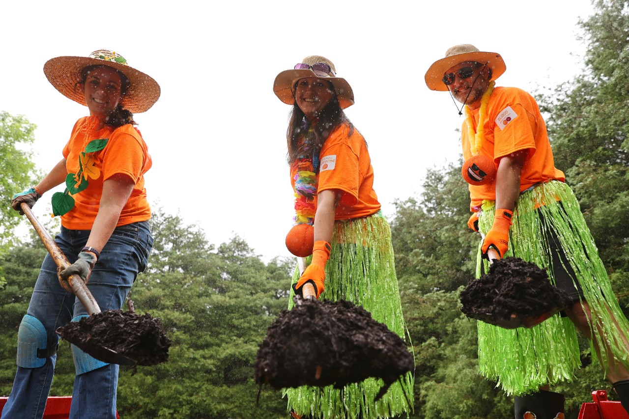 Three friends in matching shirts and hats smile for the camera while shoveling dirt in a garden.