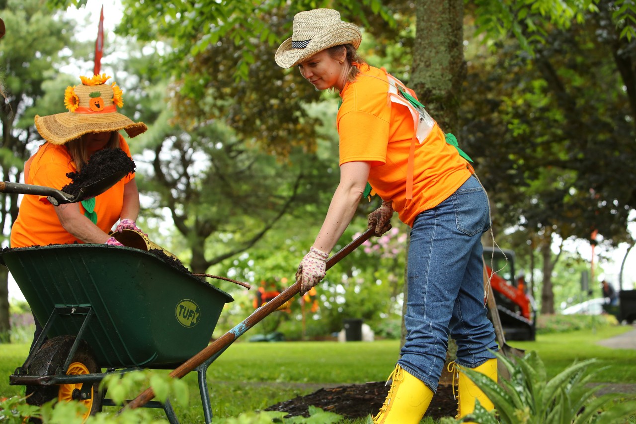 A woman wearing yellow rubber boots, an orange t-shirt, and straw hat shovels dirt from a green wheelbarrow.