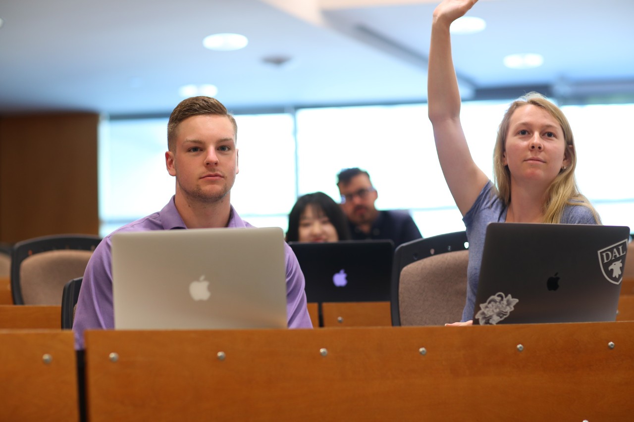 Two students sit with their laptops in a Dal lecture theatre. The student on the right is raising her hand.