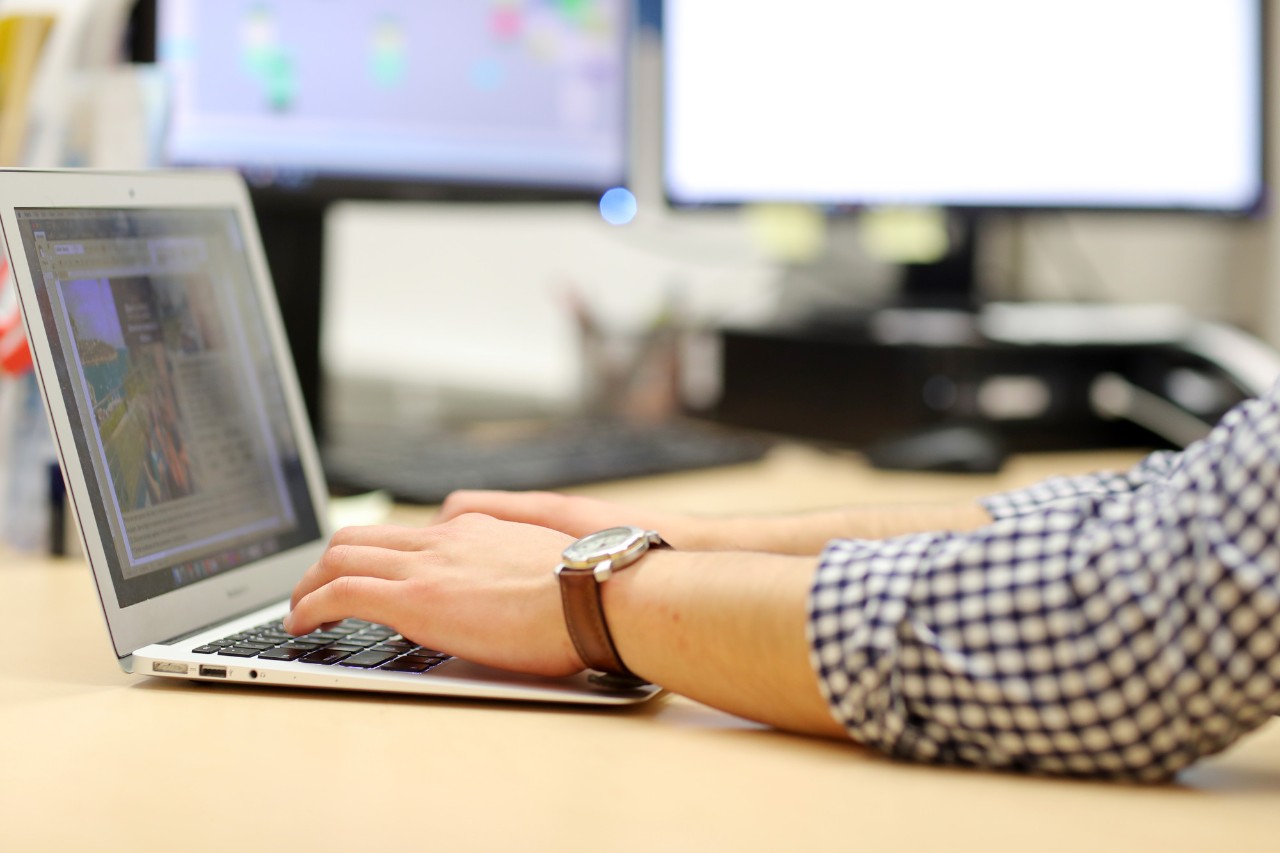 Close up of a person wearing a checked shirt and a brown watch typing on a laptop.