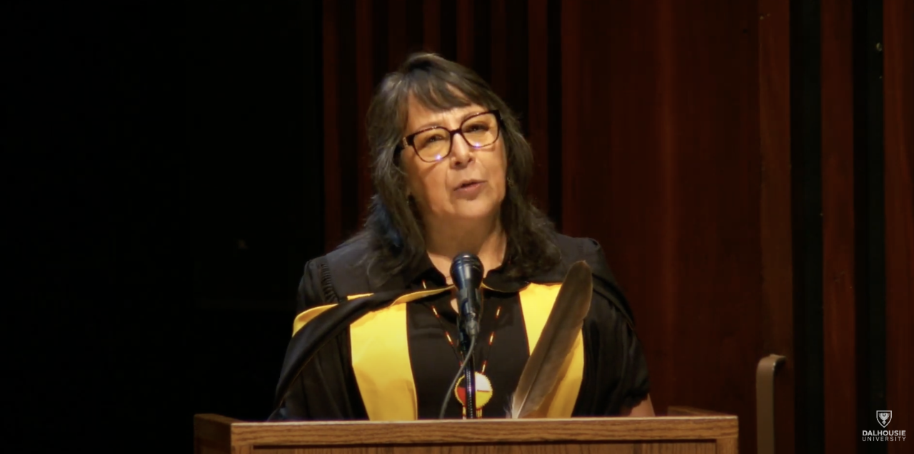 Person wearing academic dress with yellow sash speaks from lectern.