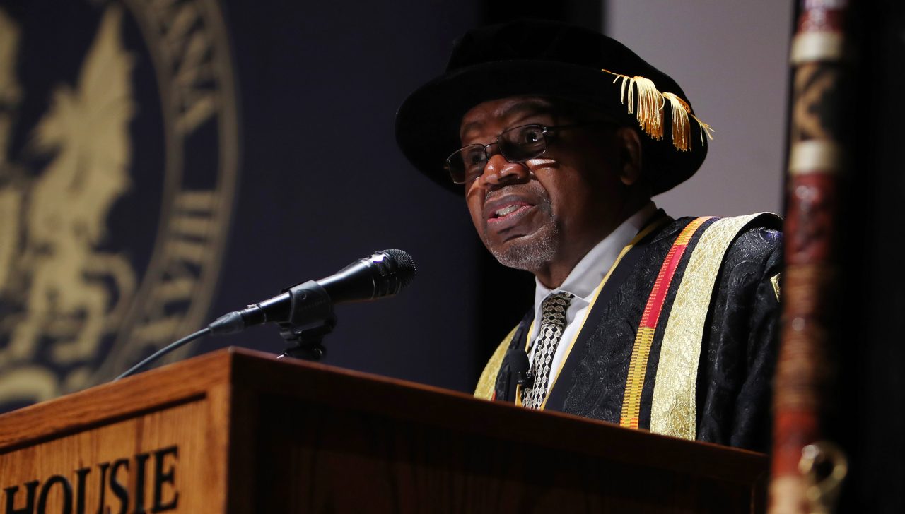 Person wearing academic robes and hat speaks from a lectern.