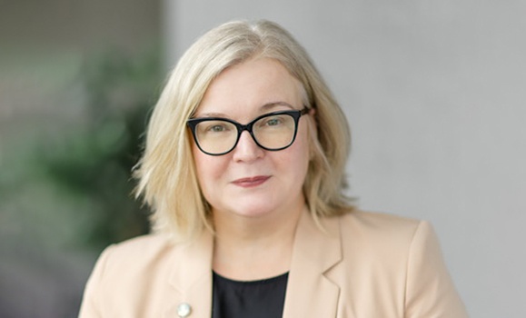 A headshot of Cathie O'Toole. She is wearing a cream blazer and wearing black framed glasses.