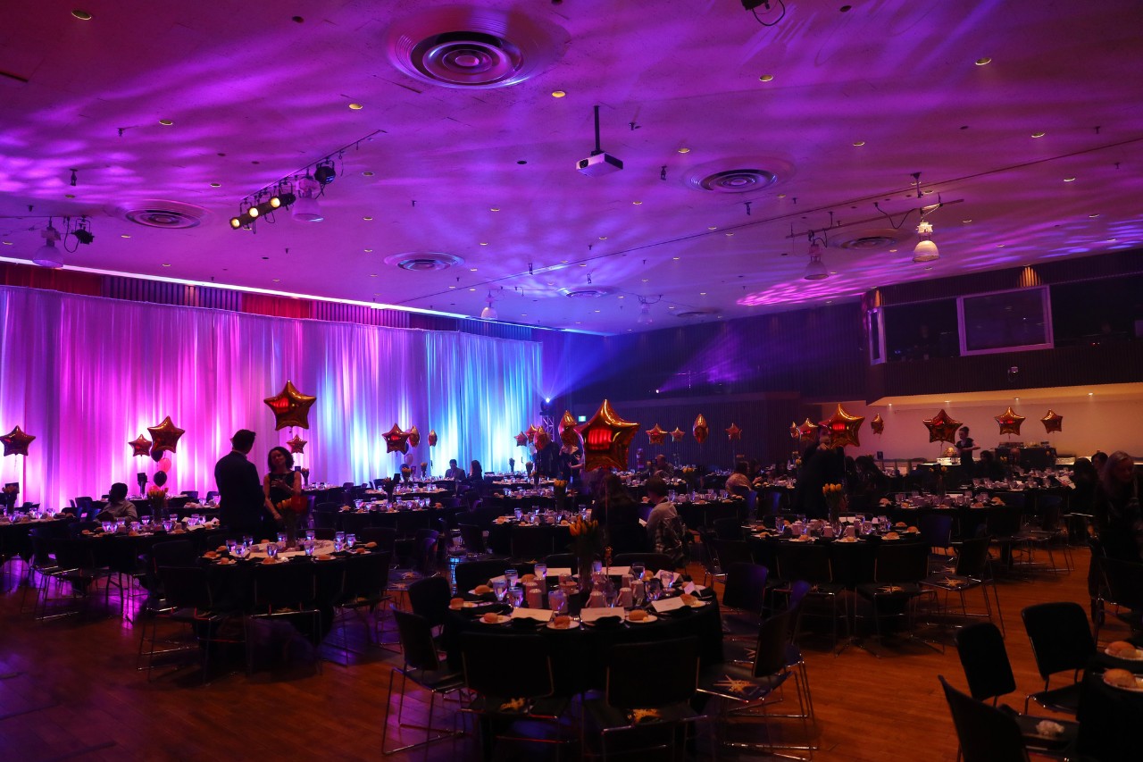 A ballroom with round tables decorated for a banquet is lit with purple and blue lighting.