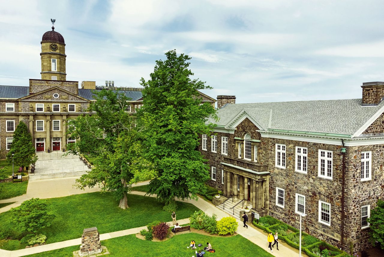 Students sitting and walking across green quad, with stone academic buildings in the background.