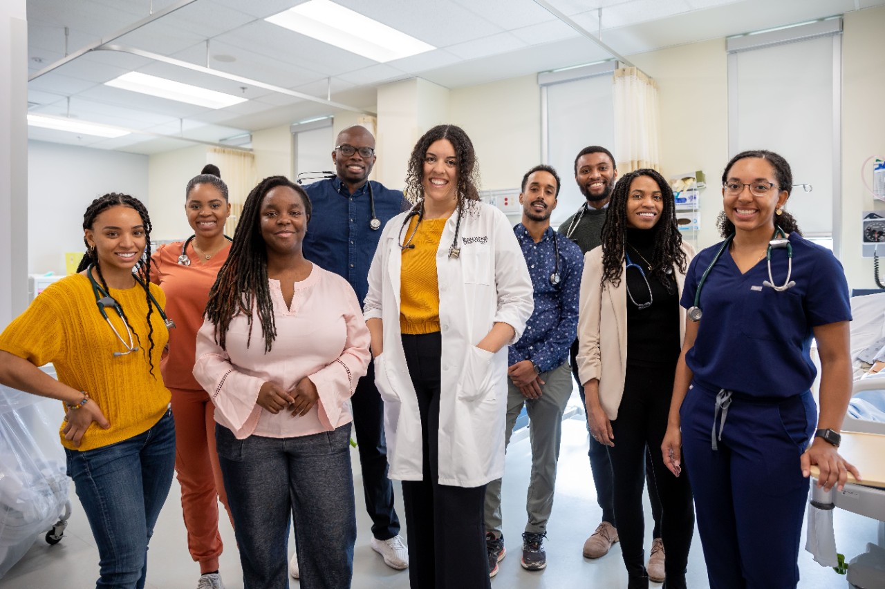 A group of health students in scrubs stand with their instructor who is wearing a white lab coat. 